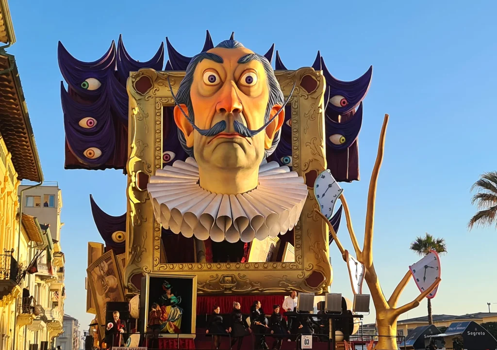 Un char du Carnaval de Viareggio avec un grand visage à la moustache retroussée dans un cadre doré, entre les immeubles du front de mer