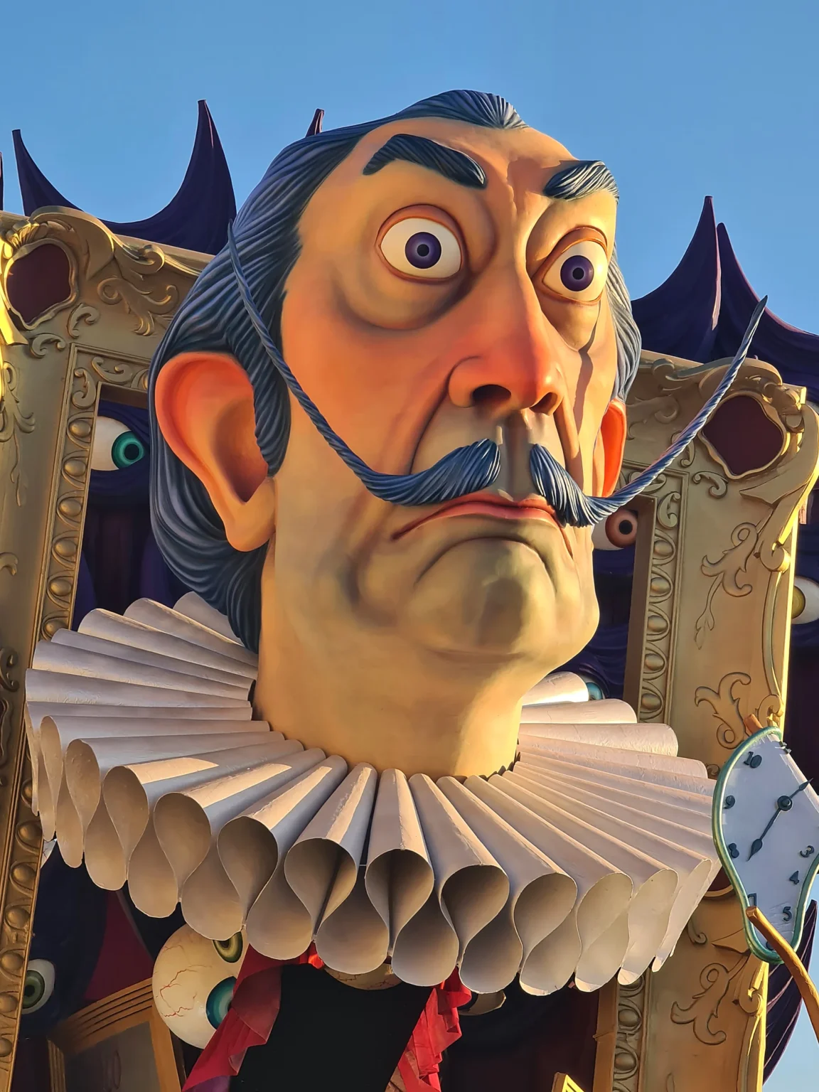 Le grand visage en papier mâché à la moustache retroussée, avec sa collerette blanche, vu de près