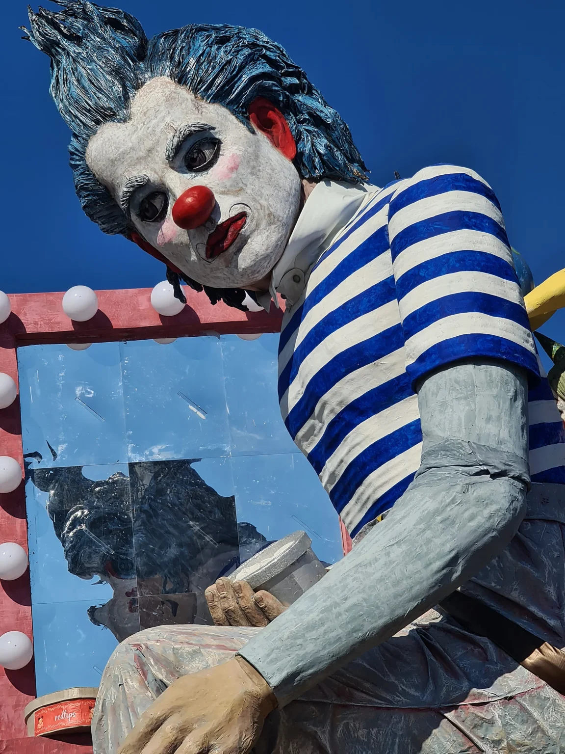 Un clown aux cheveux bleus en maillot rayé, devant un miroir à ampoules, sur un char du Carnaval de Viareggio