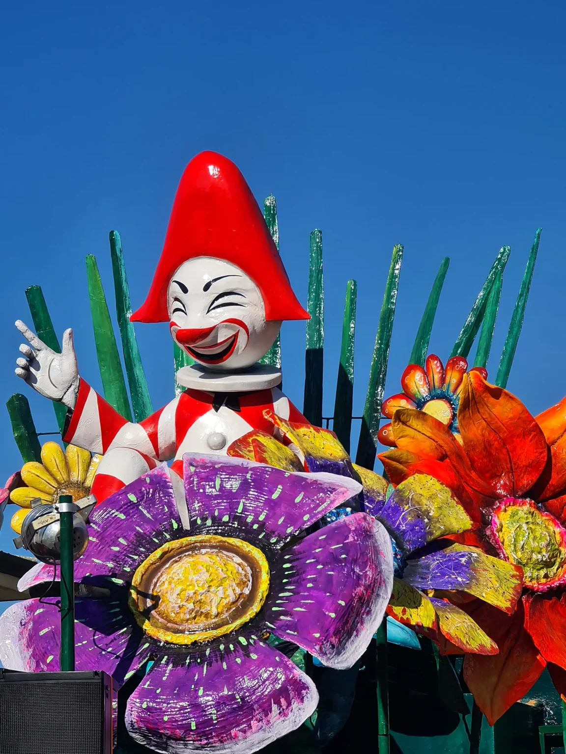 Un clown en papier mâché au chapeau rouge parmi de grandes fleurs violettes et orange, sur un char du Carnaval de Viareggio