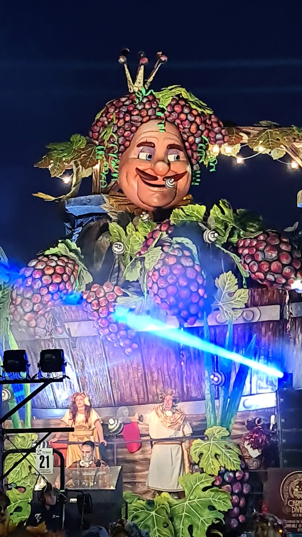 Un char du Carnaval de Viareggio illuminé le soir : un grand visage couronné de grappes de raisin